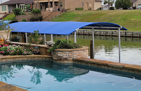 The image shows a dock canopy with a blue cover situated over a boat slip on a calm waterway. The foreground features a swimming pool surrounded by landscaping, while a residential property with a tiered yard is visible in the background. The setup emphasizes functionality and aesthetic integration of waterfront and outdoor living spaces.